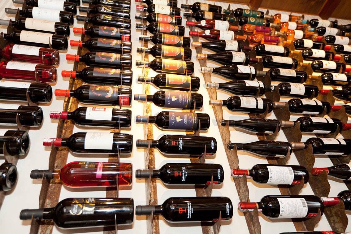 Garrafas de vinho deitadas em fileiras sobre suportes de chapa de aço com pátina, presos a uma parede clara