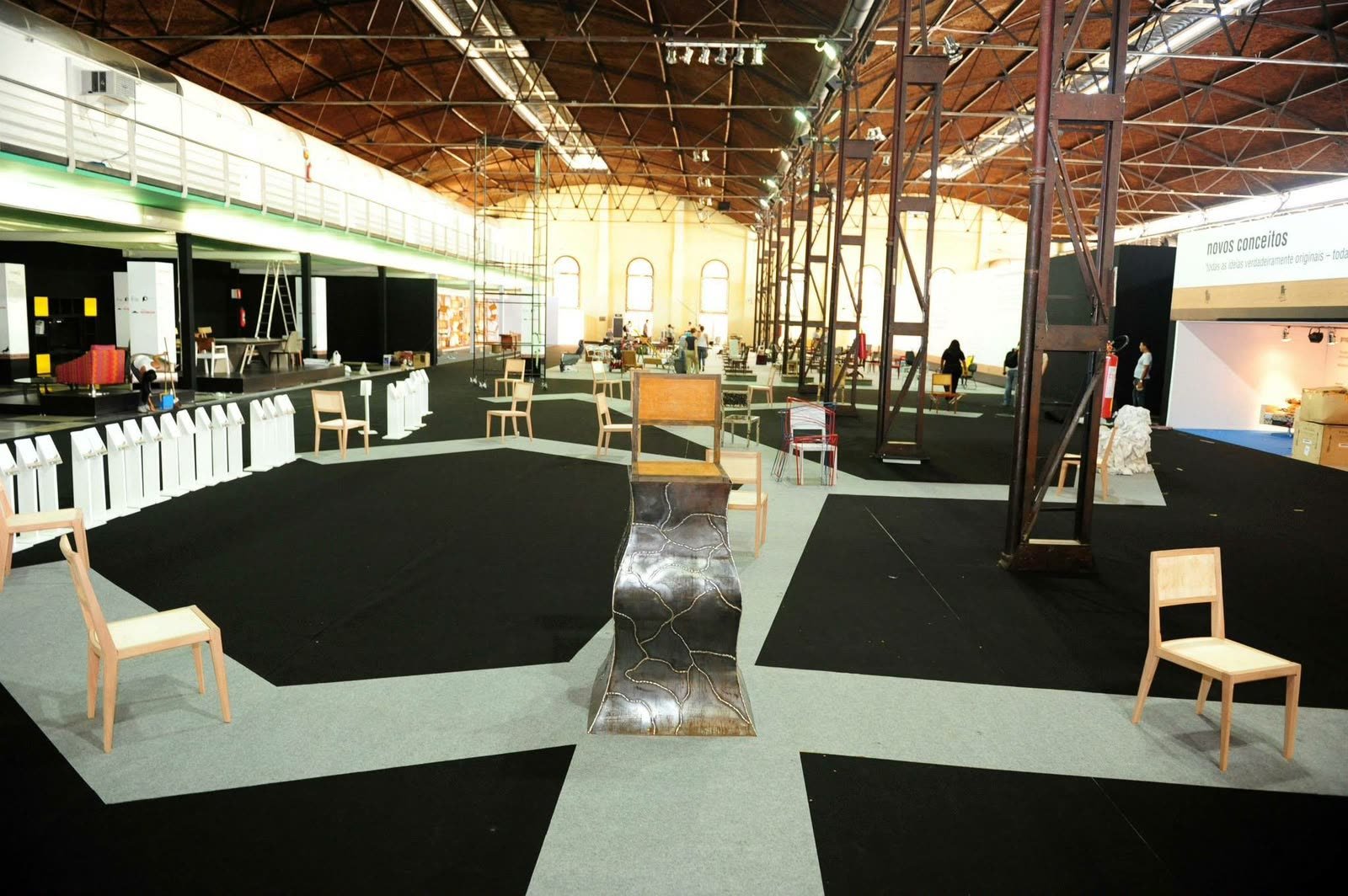 Salão de exposição com cadeiras de madeira dispostas em percursos claros sobre piso escuro e, ao centro, uma cadeira escultórica sobre base de aço