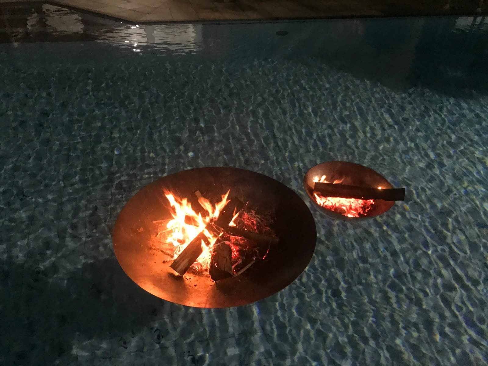 Lareiras flutuantes duplas em aço corten acesas sobre a água da piscina, à noite