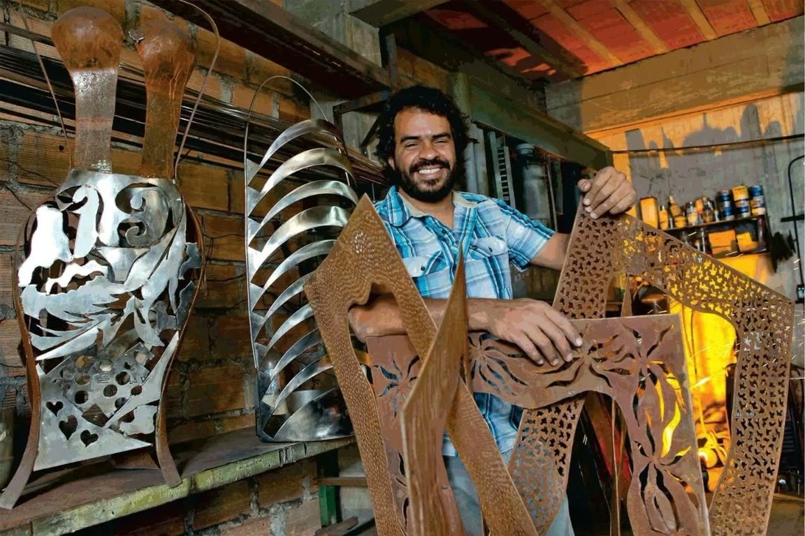 PROJETOS 32 Fabrício Carvalho, da Objetaria, sorrindo na oficina entre esculturas e painéis em aço corten e inox